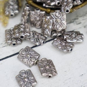 May include: Silver-colored metal beads with a rectangular shape and a floral design. The beads have a hole on each end for stringing. Some beads are scattered on a white surface, while others are in a small basket.