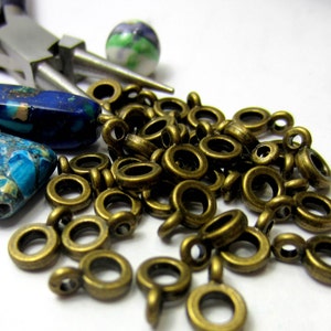 May include: A close-up of a pile of gold-colored jump rings with a small loop on one end. There are two blue and green patterned stones and a pair of silver pliers in the background.