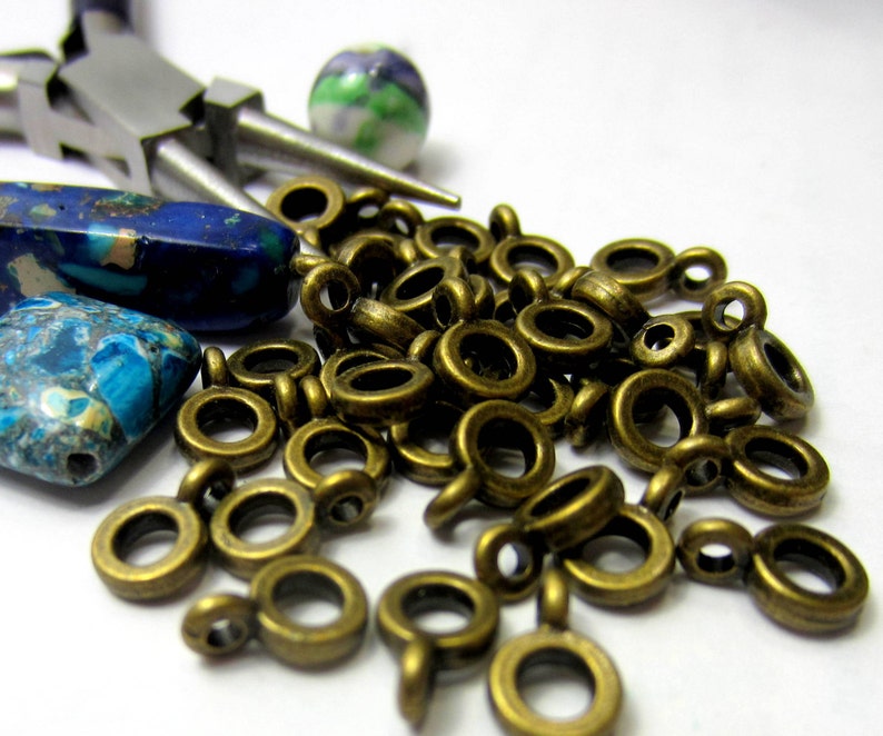 May include: A close-up of a pile of gold-colored jump rings with a small loop on one end. There are two blue and green patterned stones and a pair of silver pliers in the background.