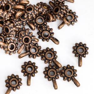 May include: Bronze-coloured metal bead caps with a floral design. Each cap has a central hole, a ring of smaller holes, and a small loop. The caps are scattered on a white surface. Ideal for jewellery making.
