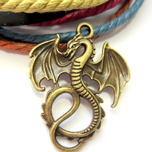 May include: A gold-toned dragon charm with intricate details, including wings and a curled tail. The charm is attached to a multi-colored braided cord.