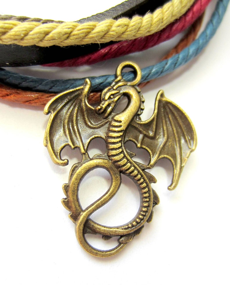 May include: A gold-toned dragon charm with intricate details, including wings and a curled tail. The charm is attached to a multi-colored braided cord.