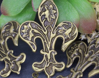 May include: Antique gold-toned metal charms in the shape of a fleur-de-lis with intricate floral designs. The charms have a textured surface and a loop at the top for attaching to jewellery or other crafts.