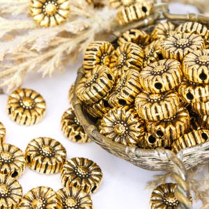以下が含まれることがあります： 花柄のデザインが施された、金色の金属ビーズのクローズアップ。ビーズは装飾的なバスケットに入っており、白い表面に散らばっています。ビーズは詳細で質感のある表面を持ち、ジュエリー作りに適しています。
