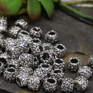 May include: A collection of silver-colored metal beads with an ornate, swirling pattern. The beads are cylindrical with a hole running through the center, and are scattered across a dark, textured surface.
