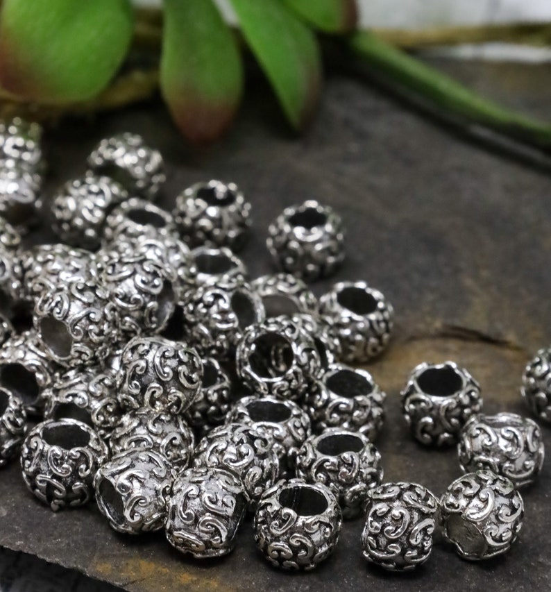 May include: A collection of silver-colored metal beads with an ornate, swirling pattern. The beads are cylindrical with a hole running through the center, and are scattered across a dark, textured surface.
