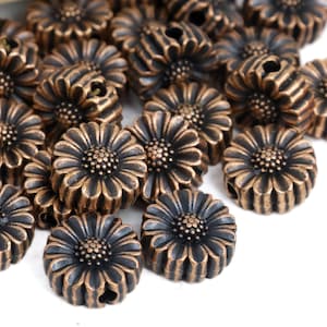 May include: A collection of bronze-coloured daisy-shaped beads. Each bead has detailed petal and centre designs, with a small hole for stringing. The beads have a metallic finish and are set against a white background.