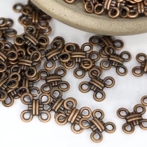 May include: A large quantity of antique gold-toned metal clasps, each shaped like a knot with four loops. The clasps are scattered on a white surface.