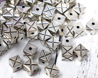 May include: A collection of silver-colored, geometric beads. Each bead is cube-shaped with a cross design on each face and a hole for stringing. The beads are scattered, suggesting their use in jewelry making or crafts.