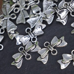 May include: Silver tone metal bow charms with two loops for attaching to jewelry or crafts. The bows are detailed with a textured surface and have a vintage look.