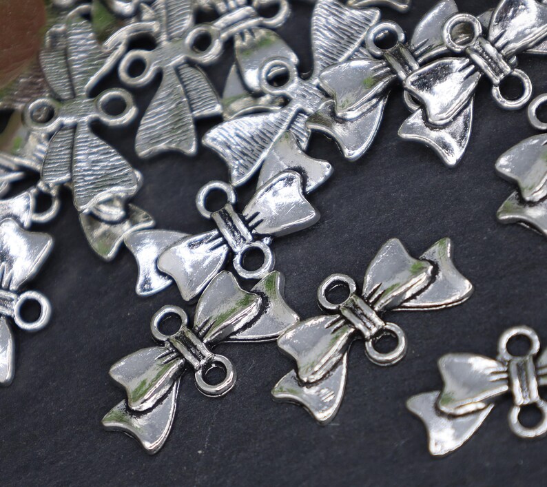 May include: Silver tone metal bow charms with two loops for attaching to jewelry or crafts. The bows are detailed with a textured surface and have a vintage look.