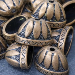 May include: A close-up of a pile of ornate, gold-toned metal caps with a black, detailed design. The caps have a rounded top and a flat bottom with a hole in the center.
