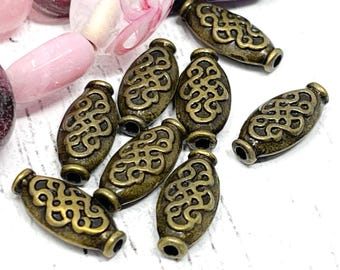 May include: Close-up of antique bronze-colored oval beads with an intricate Celtic knot design. The beads have a cylindrical shape with a hole at each end. Pink and purple beads are visible in the background.