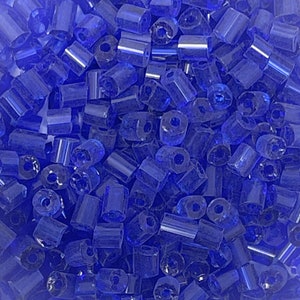 May include: A close-up view of a pile of small, blue glass beads. The beads are cylindrical in shape and have a slightly iridescent finish.