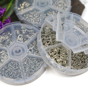 May include: Three clear plastic containers with multiple compartments, each filled with silver jump rings. The containers are round and have a hexagonal center.