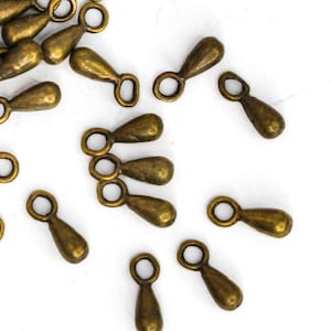 May include: A collection of small, antique brass-colored teardrop-shaped charms with a loop at the top. These jewelry findings are scattered on a white surface, ideal for crafting and jewelry making.