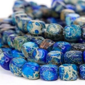 May include: Close-up of a strand of blue and teal gemstone beads. The beads are square-shaped with a polished finish, showcasing a mix of colors and patterns. The beads are strung together, creating a textured and colorful accessory.