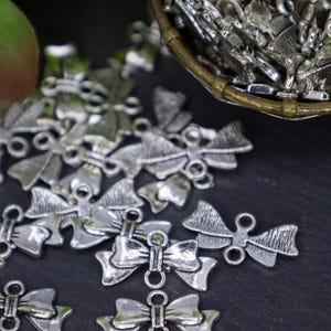 May include: Silver-toned metal bow charms with a loop on each side. The charms are scattered on a black surface.