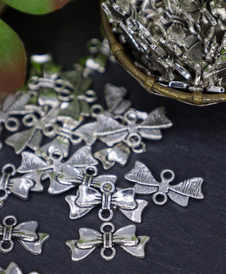 May include: Silver-toned metal bow charms with a loop on each side. The charms are scattered on a black surface.