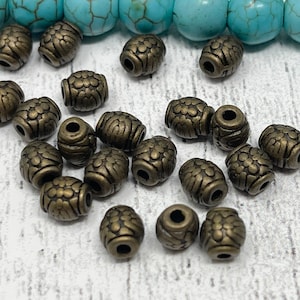 May include: A collection of 20 oval-shaped metal beads with a decorative floral pattern. The beads are a dark bronze color and have a hole in the center for stringing.