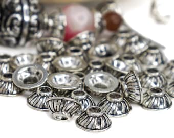 Op de afbeelding: Close-up van een verzameling zilverkleurige metalen kralenkapjes. De kralenkapjes hebben een gegroefde, klokvormige vorm met een cirkelvormige opening. Enkele andere kralen zijn zichtbaar op de achtergrond, tegen een witte achtergrond.