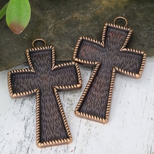 May include: Two bronze-coloured cross pendants with a textured surface and a decorative rope-like border. Each cross has a small loop at the top for hanging. The crosses are displayed on a natural background.