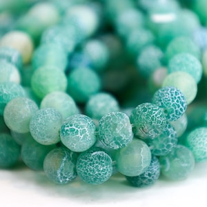 May include: Close-up of a strand of round, green beads. The beads vary in shades of green, from light to dark, with a textured, crackled appearance. The beads are strung together, suggesting they are for jewelry making or crafting.