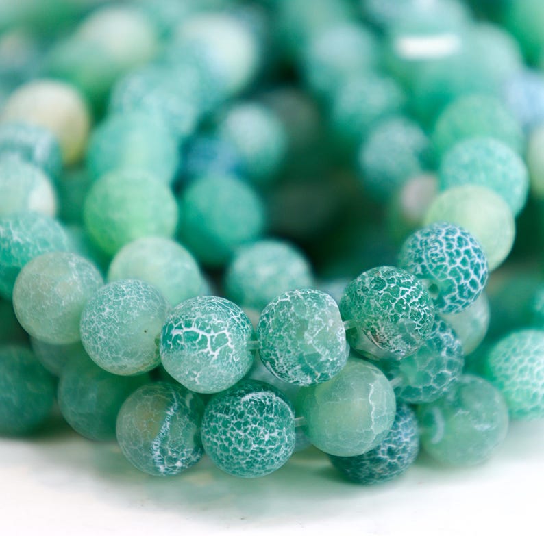 May include: Close-up of a strand of round, green beads. The beads vary in shades of green, from light to dark, with a textured, crackled appearance. The beads are strung together, suggesting they are for jewelry making or crafting.