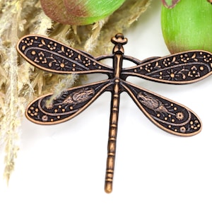May include: A copper-colored dragonfly charm with intricate floral and geometric designs. The dragonfly has a long, slender body and delicate wings.