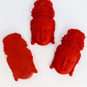 May include: Three red carved sculptures of serene faces with ornate headdresses. The sculptures are likely of a religious figure, possibly Buddha or a similar deity. The carvings are detailed and the red color is vibrant.