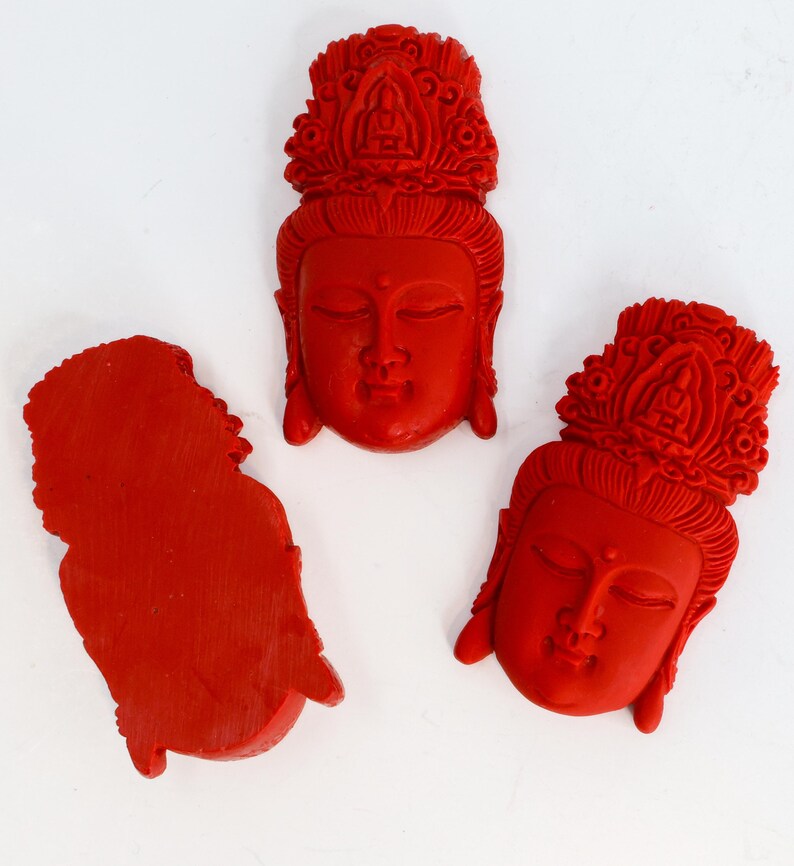 May include: Three red carved sculptures of serene faces with ornate headdresses. The sculptures are likely of a religious figure, possibly Buddha or a similar deity. The carvings are detailed and the red color is vibrant.