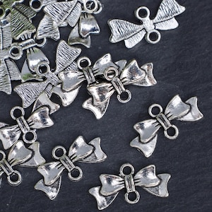 May include: A group of silver-toned metal bow charms with two loops on the back. The charms are scattered on a dark gray surface.
