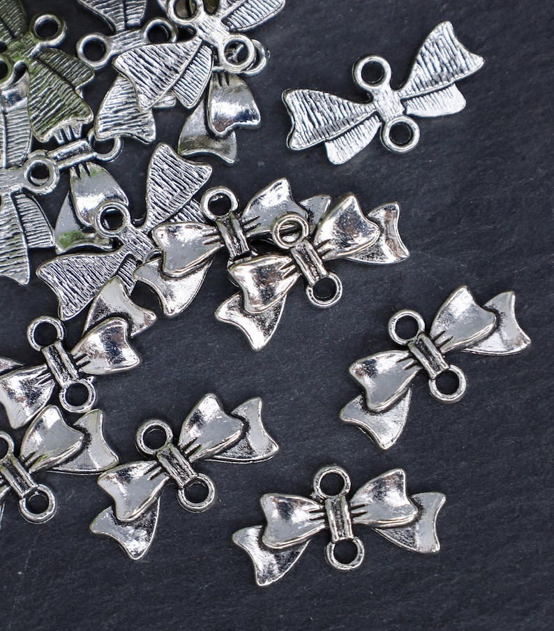 May include: A group of silver-toned metal bow charms with two loops on the back. The charms are scattered on a dark gray surface.