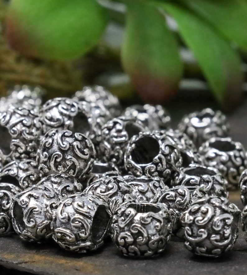 May include: A collection of silver-colored, round beads with an ornate, swirling pattern. The beads have a hollow center and are clustered together, showcasing their detailed design. The background features blurred green foliage.