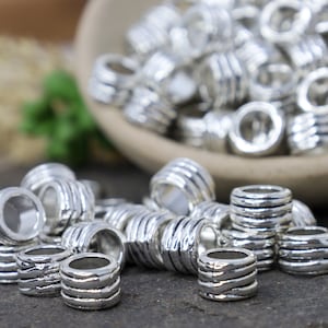 May include: A pile of silver metal beads with a textured, ridged design. The beads are arranged in a random pattern on a dark gray surface.