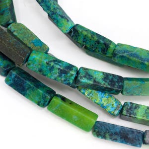 May include: Close-up of a strand of rectangular beads in shades of green and blue. The beads have a natural, textured appearance, with variations in color and pattern. The beads are strung together, creating a necklace or bracelet.
