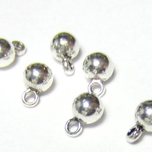 May include: Close-up of several small, shiny silver ball beads with attached loops. These jewelry-making components are scattered on a white surface, showcasing their reflective quality and simple design. Ideal for crafting earrings, necklaces, or bracelets.