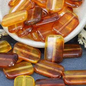 May include: A bowl filled with amber glass beads, with more beads scattered on a dark gray surface. The beads are rectangular and have a translucent, striated appearance.