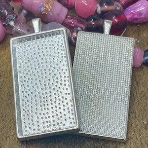 May include: Two silver-toned rectangular pendant trays with a textured surface. The trays have a loop at the top for attaching a chain or cord.