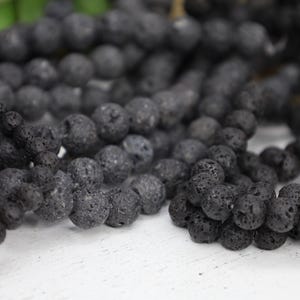 May include: A close-up of a strand of black lava rock beads. The beads are round and have a rough, textured surface.