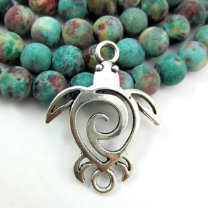 May include: A silver-colored turtle pendant with a spiral design, displayed in front of a strand of turquoise and maroon-colored beads. The pendant has a loop at the top for stringing. The beads have a matte finish.