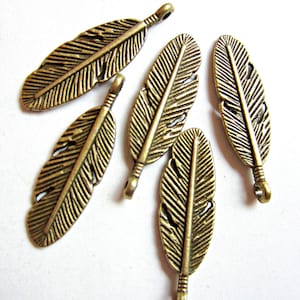 May include: Five antique bronze-colored feather charms. Each charm has a detailed feather design with a small loop at the top for attaching to jewelry or other crafts. The feathers are arranged on a white background.