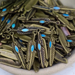 May include: A pile of antique gold-toned metal feather charms with turquoise blue enamel accents. The charms have a loop at the top for attaching to jewelry or other crafts.