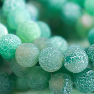 May include: Close-up of a collection of green gemstone beads. The beads are round with a cracked, textured surface, and vary in shades from light to dark green. The beads are likely used for jewelry making.