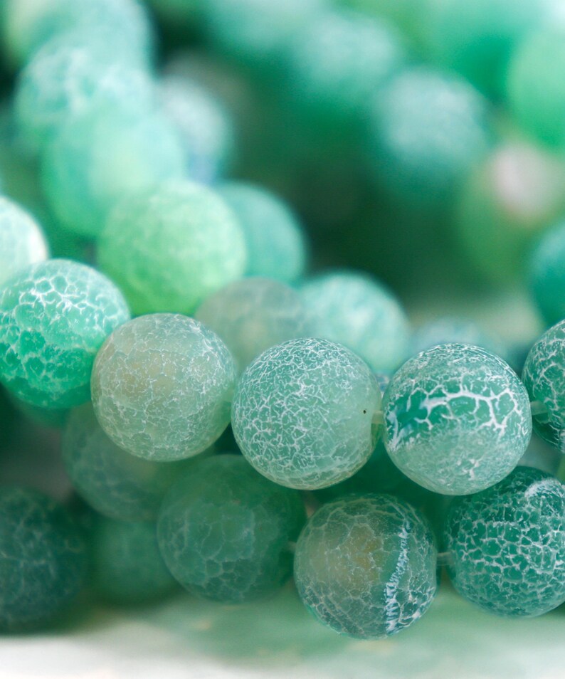 May include: Close-up of a collection of green gemstone beads. The beads are round with a cracked, textured surface, and vary in shades from light to dark green. The beads are likely used for jewelry making.