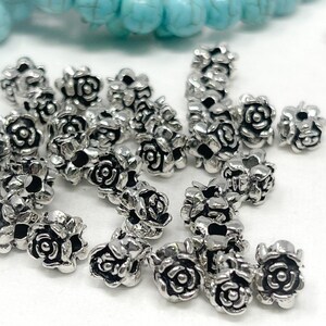 May include: A collection of small, silver-coloured rose-shaped beads. The beads are clustered together, showcasing their detailed floral design. Turquoise beads are visible in the background, adding a contrasting colour element.