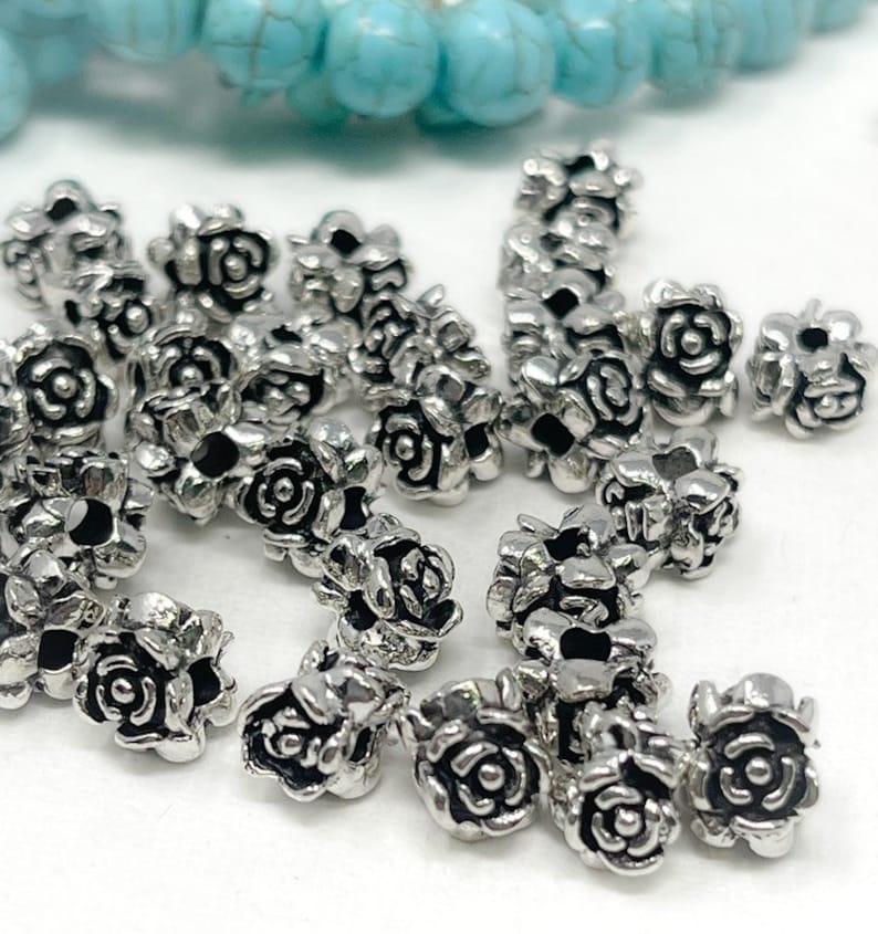 May include: A collection of small, silver-coloured rose-shaped beads. The beads are clustered together, showcasing their detailed floral design. Turquoise beads are visible in the background, adding a contrasting colour element.
