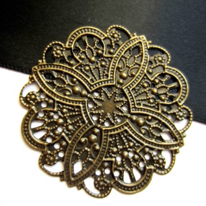 May include: Ornate, antique brass-colored filigree flower. The detailed metalwork features a central starburst design surrounded by intricate patterns and openwork. The flower is set against a black background and a white surface.
