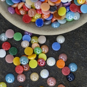May include: A collection of colorful, flat, round glass beads in various shades of red, orange, yellow, green, blue, and purple. Some beads are scattered on a dark, textured surface, while others are in a beige bowl.