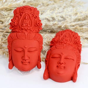 May include: Two red Buddha head carvings with closed eyes and ornate headdresses. The carvings are detailed and have a smooth finish. The background is a mix of white and beige, with some dried plants visible.